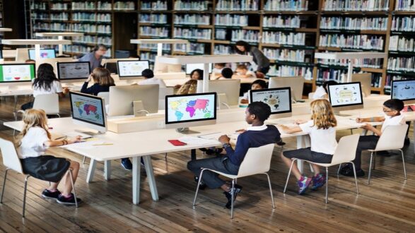 طلاب يستخدمون أجهزة الكمبيوتر في مكتبة، مع شاشات تعرض خرائط ومعلومات تعليمية. يركز الأطفال على دراستهم في بيئة تعليمية منظمة. خلفهم رفوف مليئة بالكتب.