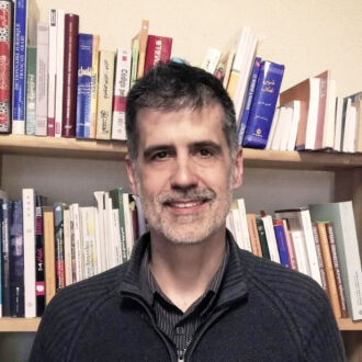 رجل مبتسم يقف أمام رف مليء بالكتب المتنوعة، يظهر في الخلفية مجموعة من الكتب باللغة العربية، تعكس اهتمامه بالقراءة والثقافة.