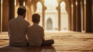 A father and son sit together on a carpeted floor in a mosque, gazing towards a sunlit entrance. The warm light creates a serene atmosphere, highlighting the bond between them.
