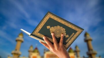 Hand holding a Quran against a blue sky and mosque backdrop, showcasing Islamic architecture and spirituality.