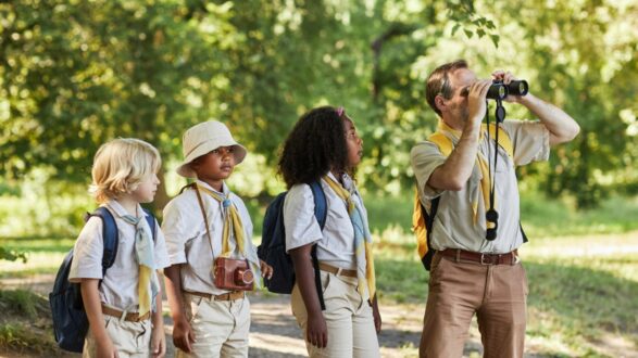 مجموعة من الأطفال يرتدون ملابس كاكي مع حقيبة ظهر، يقفون في الطبيعة بينما يستخدم أحد البالغين المنظار لمراقبة شيء ما. يبدون فضوليين ومهتمين بالبيئة المحيطة.