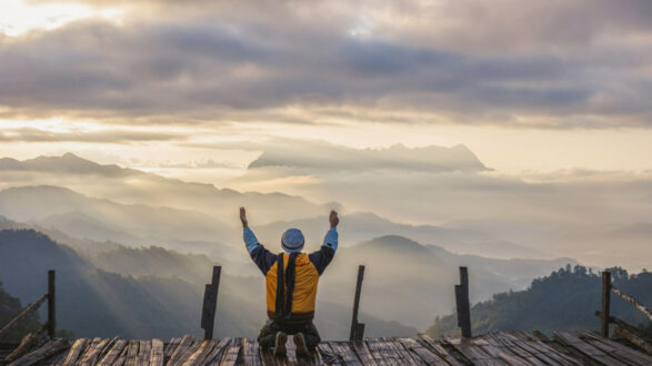 رجل يجلس على منصة خشبية مرتفعة في الجبال، يرفع ذراعيه نحو السماء. تُحيط به مناظر طبيعية خلابة للجبال المغطاة بالسحاب، مع أشعة الشمس تتسلل عبر الغيوم، مما يخلق جوًا من السكينة والإلهام.