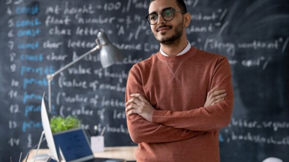رجل يرتدي سترة برتقالية يقف بفخر أمام سبورة تحتوي على معادلات رياضية وأكواد برمجة. في الخلفية، يوجد مكتب مع جهاز كمبيوتر محمول ومصباح مكتب، مما يعكس بيئة تعليمية أو عمل تكنولوجي.