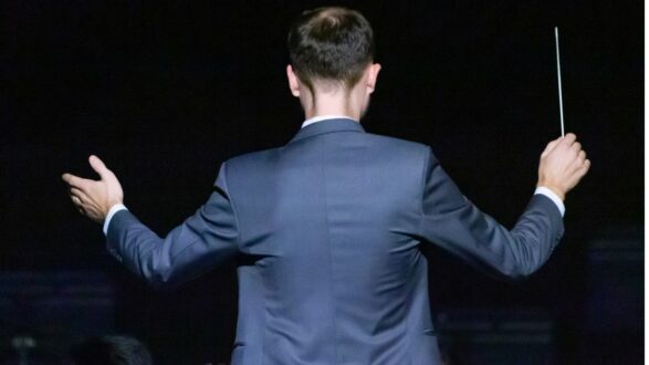 رجل يرتدي بدلة رسمية يقود الأوركسترا، يظهر من الخلف وهو يرفع عصا القيادة بيد واحدة، مع خلفية مظلمة تبرز الحركة والطاقة في الأداء الموسيقي.