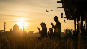 عائلة تؤدي الصلاة في حقل من القمح عند غروب الشمس، مع معالم مسجد في الخلفية. السماء مغطاة بألوان دافئة، والطيور تحلق في الأفق. تعكس الصورة أجواء الروحانية والوحدة العائلية في مشهد طبيعي.
