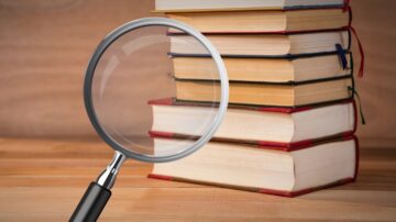 عدسة مكبرة تركز على مجموعة من الكتب المكدسة، مع تصميم خشبي خلفية، تعكس فكرة البحث والاستكشاف في عالم القراءة والمعرفة.