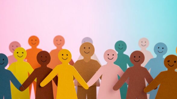 مجموعة من شخصيات ورقية ملونة تمثل التنوع والتعاون، مع خلفية ملونة تتدرج بين الوردي والأزرق. تعبر الصورة عن الوحدة والشمولية من خلال الأشكال السعيدة والمختلفة.