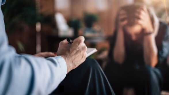 رجل يجلس ويكتب في دفتر ملاحظات بينما يبدو شخص آخر في الخلفية مضغوطًا ويضع يديه على رأسه. تعبيرات الوجه توضح القلق أو الإجهاد. تبرز الأجواء الداخلية للمكان مع النباتات والخلفية الدافئة.