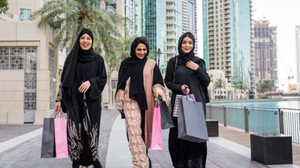 نساء يرتدين العباءات التقليدية يتسوقن في منطقة حضرية حديثة، يحملن أكياس تسوق ملونة، مع ناطحات سحاب وأشجار النخيل في الخلفية.