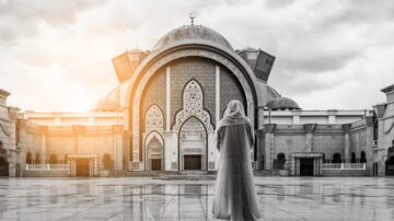 امرأة ترتدي الحجاب وتقف أمام مسجد كبير بتصميم معماري مميز، مع سماء غائمة وأشعة شمس تتسلل، تعكس الأجواء الروحانية والجمال الثقافي.