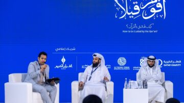 مناقشة حول كيفية التوجيه وفقًا للقرآن في حدث يجمع متحدثين من خلفيات مختلفة. يظهر شخصان يجلسان على منصة، أحدهما يرتدي الزي التقليدي القطري، والآخر يرتدي بدلة. يتحدث أحدهم بينما يستمع الآخر. الخلفية زرقاء مع شعارات رعاة الحدث.