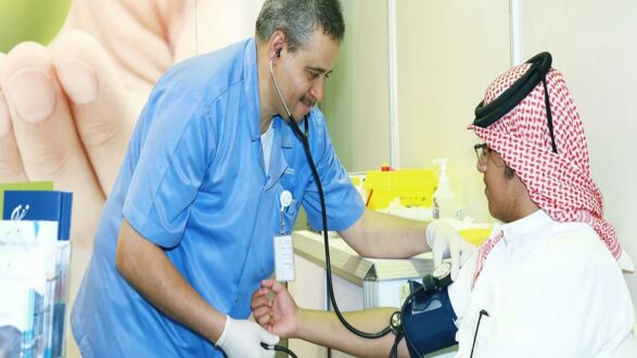 فحص ضغط الدم لمريض يرتدي الزي السعودي التقليدي، مع طبيب يستمع إلى نبضات قلبه باستخدام سماعة طبية. الخلفية تظهر يداً تمسك بيد طفل، مما يوحي بالاهتمام بالرعاية الصحية.