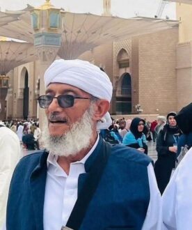 رجل مسن يرتدي غطاء رأس أبيض ونظارات شمسية، يقف في ساحة مزدحمة بمكة المكرمة، حيث يظهر المبنى التاريخي خلفه. الحشد يضم زوارًا يرتدون ملابس تقليدية.