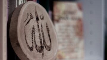 ختم خشبي مزخرف برمز إسلامي، موضوع على رف مليء بالكتب. يظهر في الخلفية نص مطبوع بألوان دافئة.