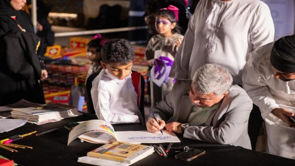 رجل مسن يجلس على طاولة ويكتب بحماس بينما يشاهد صبي صغير. مجموعة من الأشخاص، بينهم أطفال يرتدون ملابس ملونة، تتجمع حولهم في حدث ثقافي أو تعليمي. الكتب والأدوات الفنية متناثرة على الطاولة، مما يشير إلى نشاط إبداعي.