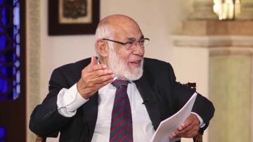 رجل مسن ذو لحية بيضاء يرتدي بدلة سوداء وقميص أبيض مع ربطة عنق ملونة، يجلس في مكان داخلي يتحدث بحماس، ممسكًا بورقة، ويظهر تعبير وجهه مفعم بالحيوية.