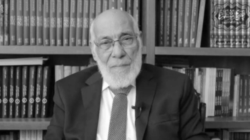 رجل مسن ذو لحية بيضاء يرتدي بدلة داكنة وربطة عنق، يجلس أمام مكتبة مملوءة بالكتب. يظهر في الخلفية أرفف تحتوي على مجموعة متنوعة من الكتب، مما يعكس أجواء أكاديمية.