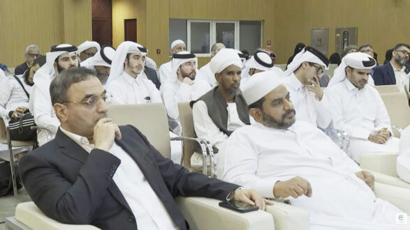 رجال ونساء يرتدون ملابس تقليدية في قاعة محاضرات، بعضهم يجلس على كراسي بينما يركزون على المتحدث. يظهر الحضور في بيئة رسمية، مع تنوع في الأزياء، مما يعكس ثقافة المجتمع.