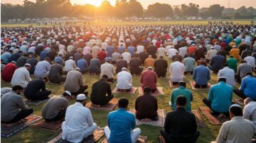 A large group of people in traditional attire is gathered outdoors, participating in a prayer at sunrise. The scene captures a diverse assembly of individuals seated on prayer mats, with tents in the background. The soft light of dawn illuminates the gathering, creating a serene atmosphere for this communal worship event.