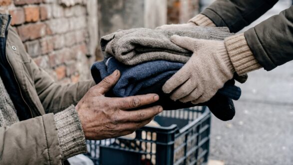 رجل يقدم مجموعة من الملابس الدافئة لرجل آخر يجلس على الرصيف، مع خلفية جدران من الطوب. تُظهر الصورة أهمية العطاء والمساعدة في المجتمع.