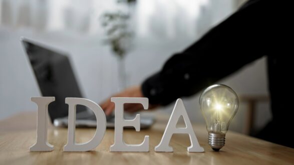 إضاءة مصباح مع كلمة "IDEA" على طاولة خشبية، وخلفية تظهر شخصًا يعمل على كمبيوتر محمول. تعكس الصورة مفهوم الإبداع والتفكير.
