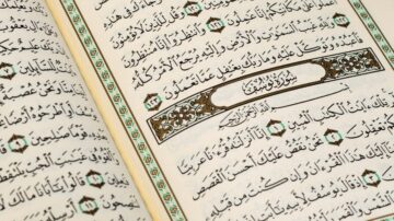 كتاب القرآن الكريم مفتوح على صفحات تتضمن آيات مكتوبة بخط عربي جميل، مع تزيينات نباتية ملونة حول النص. يبرز النص بشكل واضح، مما يعكس جمال الخط العربي.