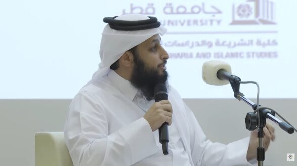 رجل يرتدي الزي التقليدي العربي يتحدث في حدث أكاديمي، مع وجود شعار جامعة قطر في الخلفية. يظهر المتحدث وهو يتفاعل مع الجمهور من خلال الميكروفون.