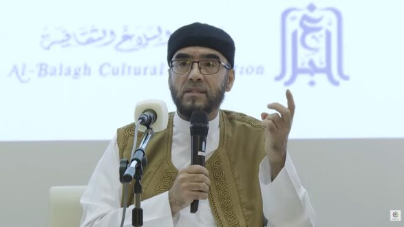 رجل يرتدي ملابس تقليدية ويضع نظارات، يتحدث أثناء ندوة ثقافية. خلفه شعار مؤسسة البلاغ الثقافية. يركز على تقديم أفكار مهمة للجمهور.