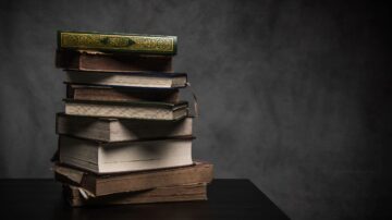 كتب مكدسة بشكل عشوائي على طاولة، مع إضاءة خافتة تبرز تفاصيلها. الكتاب العلوي له غلاف مزخرف باللون الأخضر، بينما الكتب الأخرى تتنوع في الألوان والأحجام. الخلفية مظلمة، مما يضيف عمقاً للمشهد.