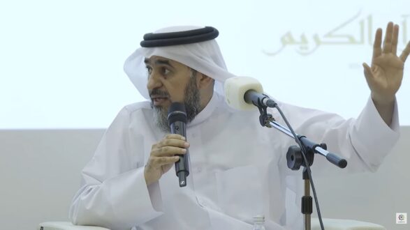 رجل يرتدي الزي التقليدي العربي، يتحدث في ندوة باستخدام ميكروفون، مع وضع يده في الهواء، وخلفه لافتة تحمل نصًا باللغة العربية.