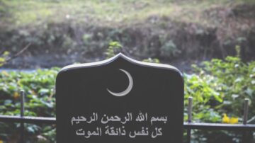 نقش على لوحة حجرية تحتوي على نص عربي يقرأ "بسم الله الرحمن الرحيم كل نفس ذائقة الموت" مع رسم هلال، محاط بنباتات خضراء وخلفية طبيعية.