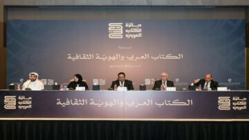 ندوة حول الكتاب العربي والهوية الثقافية، تضم متحدثين يجلسون على منصة في مؤتمر. يظهر في الصورة عدة أشخاص يرتدون ملابس رسمية، مع وجود لافتات تحمل عنوان المؤتمر، وخلفية تتضمن شعارات ثقافية.