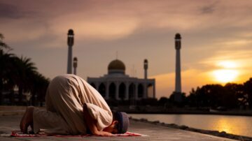 A person performing a prayer in front of a mosque during sunset, with palm trees in the foreground and minarets visible in the background. The sky is filled with warm hues of orange and purple, creating a serene atmosphere.