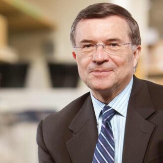 رجل مسن يرتدي بدلة رسمية وقميص مزخرف، يجلس مبتسمًا في بيئة مكتبية، مع خلفية ضبابية تحتوي على أرفف.