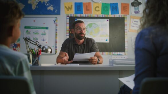 مدرس يجلس على طاولة في فصل دراسي، يتحدث إلى طالبين. خلفه لوحة ترحيب ملونة، ومخططات تعليمية على الجدران، وأدوات مكتبية مرتبة.