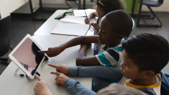 أطفال في فصل دراسي يستخدمون جهاز لوحي. يظهر طفلان يشيران إلى الشاشة بينما يركز الآخرون على الكتابة في دفاترهم. تعكس الصورة بيئة تعليمية تفاعلية تشجع على التعلم الجماعي واستخدام التكنولوجيا.