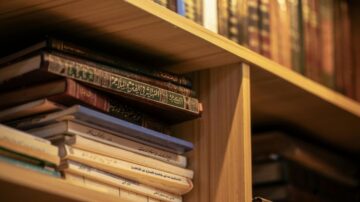 كتب عربية متنوعة مرتبة على رف خشبي، تضم عناوين مختلفة في مجالات الأدب والتاريخ. الخشب يبدو دافئاً وتفاصيل الكتب واضحة، مما يعكس أهمية المعرفة والثقافة.