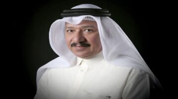 رجل يرتدي غطاء رأس تقليدي وعباءة بيضاء، يظهر بابتسامة خفيفة أمام خلفية داكنة، يعكس الأناقة والثقافة العربية.