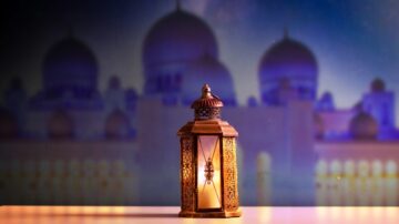 فانوس مزخرف بتصميم تقليدي مع إضاءة دافئة، متمركز أمام خلفية ضبابية تظهر قباب معمارية، مما يضفي جواً ساحراً يعكس الثقافة العربية.