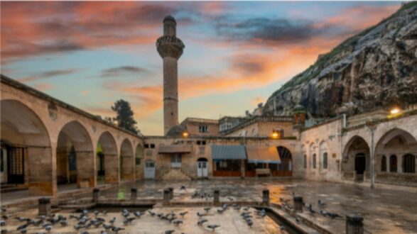 ساحة تاريخية في مدينة قديمة، تظهر فيها قبة ومسجد مع مئذنة مرتفعة. السماء تتلون بألوان الغروب، والطيور تتجمع حول بركة مياه. الواجهات الحجرية تعكس التراث المعماري الغني، مع صخور خلفية تعزز المنظر الطبيعي.