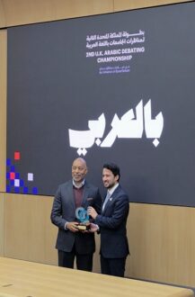 رجلان يقفان أمام شاشة كبيرة تعرض شعار بطولة المناظرات العربية في المملكة المتحدة. أحدهما يحمل جائزة بينما الآخر يبتسم، في جو احتفالي. المناسبة تبرز أهمية المناظرات باللغة العربية بين الجامعات.