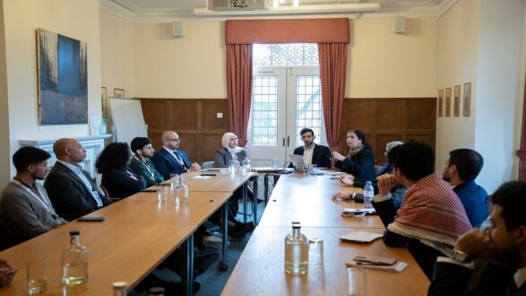 اجتماع لمجموعة من الأشخاص حول طاولة مستطيلة في غرفة ذات إضاءة جيدة. يتحدث أحد المشاركين بينما يستمع الآخرون بانتباه. توجد زجاجات مياه وكؤوس على الطاولة، وخلفهم نافذة كبيرة تسمح بدخول الضوء الطبيعي.