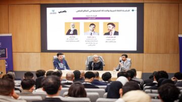 ندوة حول اللغة العربية والهوية الرقمية، تتضمن ثلاثة متحدثين في منصة أمام جمهور، مع عرض تقديمي في الخلفية يتضمن عناوين وأسماء المتحدثين. الحضور يتكون من مجموعة متنوعة من الشباب المهتمين بالثقافة واللغة.