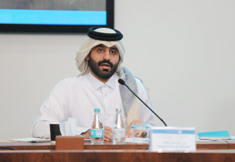 رجل يتحدث في مؤتمر، يرتدي الزي التقليدي القطري مع عمامة، يجلس أمام طاولة تحتوي على زجاجات ماء وملاحظات، خلفه شاشة عرض.