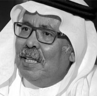 رجل مسن يرتدي الزي التقليدي العربي مع غطاء رأس، يظهر تعابير وجهه بوضوح أثناء حديثه، خلفية ضبابية.