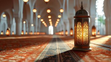 Decorative lantern illuminating a mosque interior with intricate patterns, warm lighting, and a patterned carpet. Soft bokeh effect from hanging lights in the background enhances the serene atmosphere.