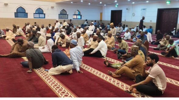 A large group of people seated on a carpeted floor inside a mosque, participating in a communal gathering or prayer. The interior features traditional architectural elements with arched windows and decorative patterns. Attendees are dressed in various cultural attire, reflecting a diverse community.