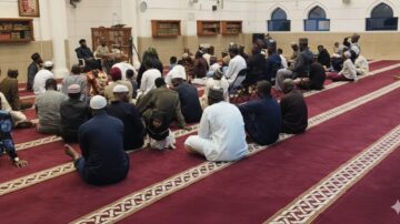 A diverse group of people seated on a red carpet in a mosque, attentively listening to a speaker at the front. The setting features Islamic architecture with arched windows and shelves of religious texts.