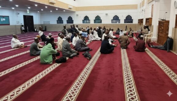 Men sitting on red carpeted floor in a mosque, engaged in a religious gathering. The setting features ornate architectural details and soft lighting, creating a serene atmosphere for worship and community.