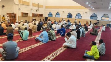 A large group of people seated in a mosque during a religious gathering, with men and children wearing traditional attire. The interior features a red carpet, decorative arches, and a speaker at the front, emphasizing community and worship.